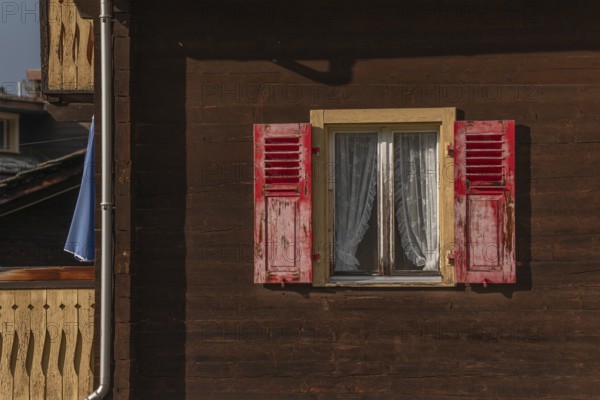 The wooden house stands in a mountain landscape, the windows are decorated with red shutters. Lace curtains add a welcoming touch to this natural atmosphere. Grachen, Viege, Valais, Switzerland