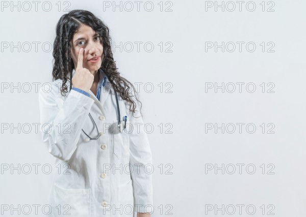 Suspicious young doctor pointing at eye, isolated. Suspicious female doctor looking you isolated
