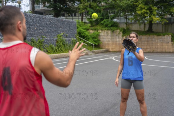 Two baseball players practicing catching on a court, with a woman wearing a glove attempting to catch a ball thrown by a man dressed in a red shirt, showcasing teamwork and skill