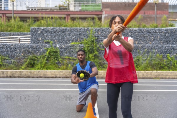 Engaging in an outdoor baseball practice, a woman swings a bat with focus while a man kneels, poised to catch the ball, showcasing teamwork and skill under the summer sun