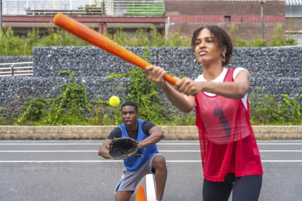 Female baseball player swinging a bat to hit a ball while a male catcher prepares to catch it. Action packed moment capturing the excitement of the game in a sunny park setting