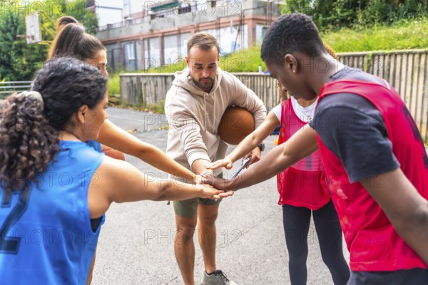 Diverse group of basketball players and their coach gather in a huddle, joining hands together, symbolizing their unity, teamwork, and sportsmanship before a game on an outdoor court