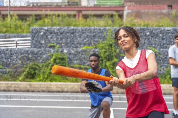 Young woman with red training shirt holding a baseball bat ready to hit the ball during a match with her team, a teammate is crouching behind her ready to catch the ball