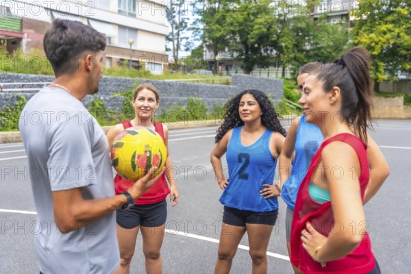Male coach holding a soccer ball, engaging in conversation with a group of female soccer players dressed in vibrant jerseys on an outdoor court, focusing on strategy and teamwork