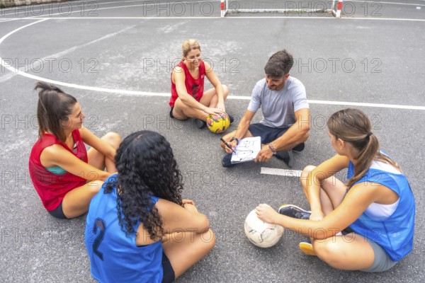 Soccer coach sitting on the field, drawing game strategy on a clipboard while female players listen attentively during training, fostering teamwork and skill development