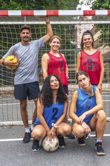 Five young soccer players, wearing numbered jerseys, are posing for a team photo on an outdoor court, holding soccer balls and standing in front of the net