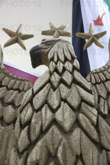 A large sculpture of Syria's national emblem eagle is displayed inside a pavilion at the Damascus International Fair following the fall of Assad's rule. Visitors stop to take photos beside the tan-coloured eagle, whose wings represent Syria's 14 governorates and tail feathers symbolize the five main regions of the country. Officially adopted on 3 July 2025, the emblem has become a marker of Syria's new visual identity, Damascus, Damascus, Syria