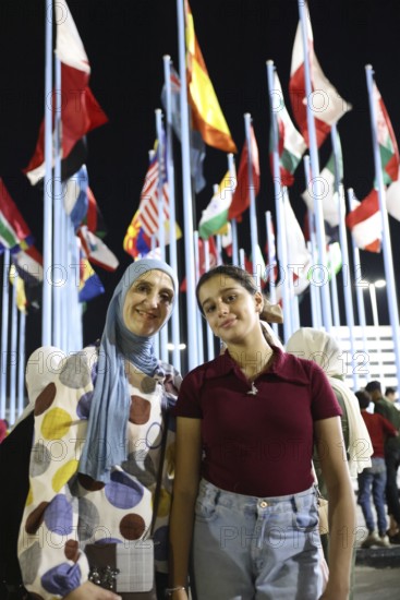 Visitors take souvenir photos at the central square of the Damascus International Fair, with the flags of 22 participating countries displayed in the background. The fair, held after the fall of Assad's regime, drew large crowds exploring cultural and economic exhibitions, Damascus, Damascus, Syria