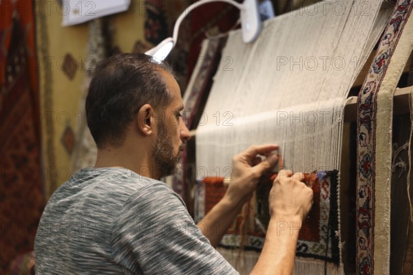 A Syrian weaver showcases the craft of traditional textile-making inside the Damascus International Fair. The artisan ties threads by hand to create a patterned carpet, reflecting Syria's centuries-old weaving heritage preserved despite years of conflict, Damascus, Damascus, Syria