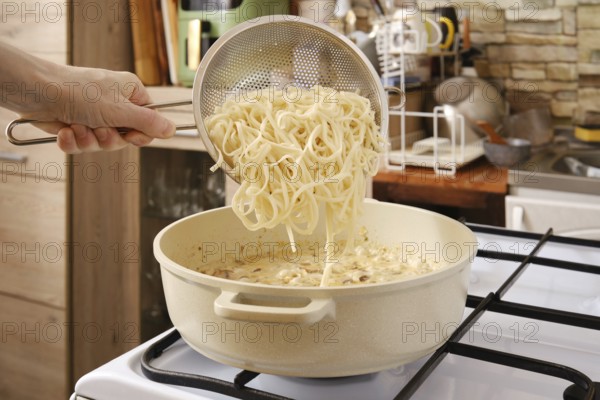 A hand adds cooked noodles from strainer in a pot of creamy mushroom sauce on the stove