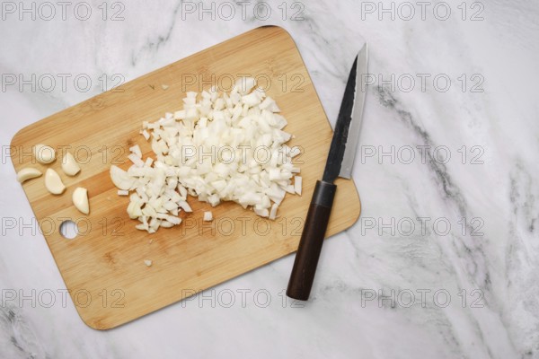 Diced onions and whole garlic cloves rest on a wooden cutting board beside a sharp knife. The countertop features a sleek, modern design, showcasing kitchen preparations with fresh ingredients