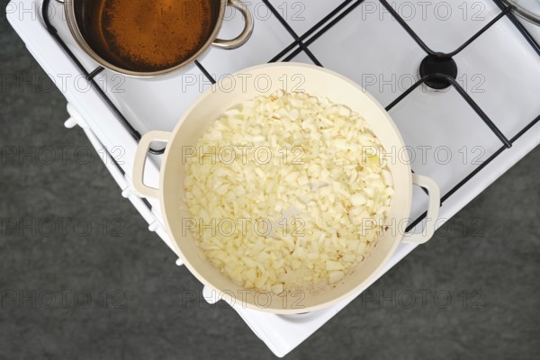 Chopped onions cooking in a white pan over medium heat on a stove. A pot of simmering broth sits close by, releasing steam into the kitchen air, suggesting a flavourful dish in preparation