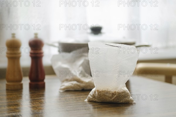 Pearl barley is prepared for cooking in convenient sachets placed on a wooden table. Nearby, salt and pepper shakers add flavor to the scene, while a pot simmers in the background