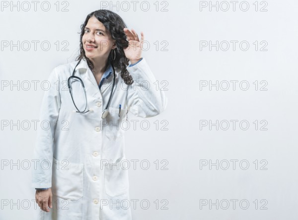 Latin female doctor listening to gossip, looking at camera. Doctor with hand on ear listening to rumor, isolated