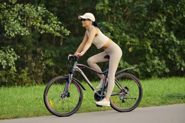 A woman enjoys riding her mountain bike along a serene path in a park. Dressed in fitted activewear, she pedals confidently while surrounded by lush greenery under a bright sun