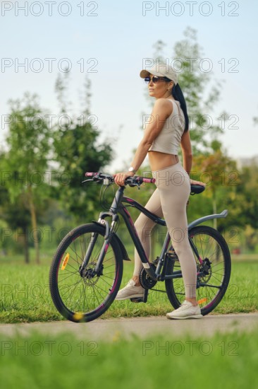 A woman stands with her bicycle in a green park, wearing athletic clothing and sunglasses. She appears confident and ready for a biking adventure in the warm sunlight