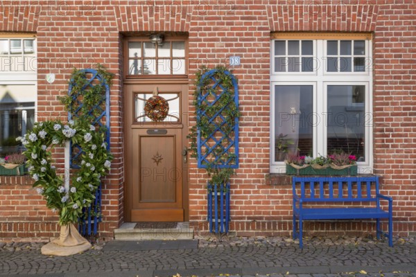 A decorated brick house façade with a blue bench and floral decorations next to the entrance, North Rhine-Westphalia, Germany