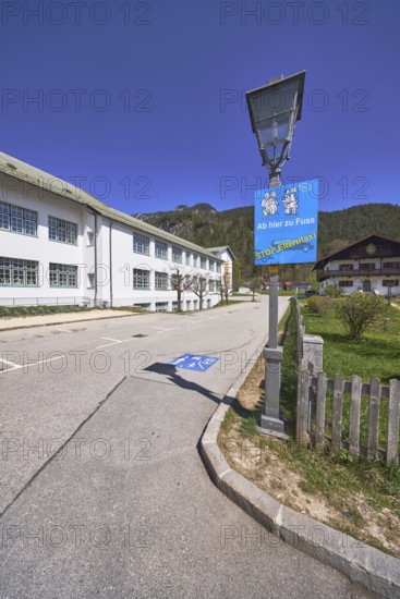Primary and secondary school Bischofswiesen, sign Stop parent taxi, building, lantern, problem parent taxi, wooden fence, hill, coniferous forest, blue sky, cirrostratus clouds, intersection Göllstraße with Schulstraße, Bischofswiesen, district Berchtesgadener Land, Bavaria, Germany