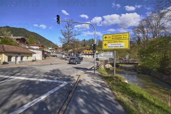 Signposts to Königssee, Berchtesgaden, Maria Gern and natural swimming pool Achauer Weiher, streets, traffic lights, buildings, trees, hills, coniferous forest, river Bischofswieser Ache, car and pedestrian bridge, blue sky, cumulus clouds, intersection between federal road 20, main road, Aschauerweiherstraße and Steingasse, Bischofswiesen, district Berchtesgadener Land, Bavaria, Germany