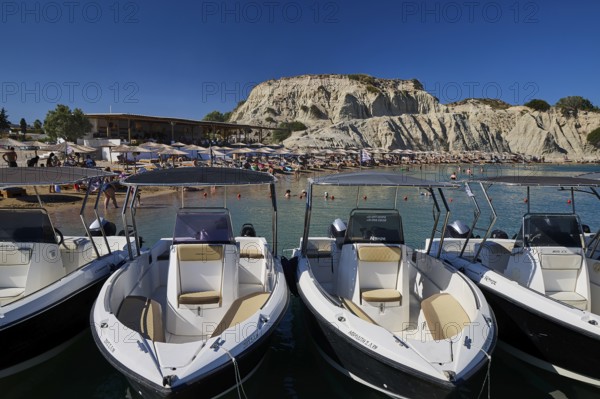 Several boats are moored on a jetty with a rocky beach in the background. Holidaymakers relaxing on the beach and in the water, Limanaki beach, sandstone cliffs, Kolymbia, Rhodes, Dodecanese, Greek islands, Greece