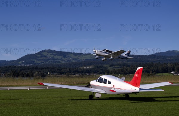 PA28 Archer II 180 HP identification aircraft registration HB-PMG during take-off over Piper Archer II HB-PPY on the ground, Wangen-Lachen airfield, LSPV, Canton Schwyz, Switzerland