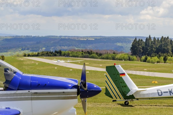 In the background runway and landing strip on it tow launching motorised aircraft towing glider glider taking off, in the foreground left nose tip of parked single-engine propeller aircraft right rudder of motor glider in parking position, Wasserkuppe airfield, Gersfeld, district of Fulda, Hesse, Germany