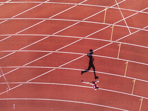 Aerial view of a female individual (runner) on a red athletics track (tartan track) with shadow as a silhouette