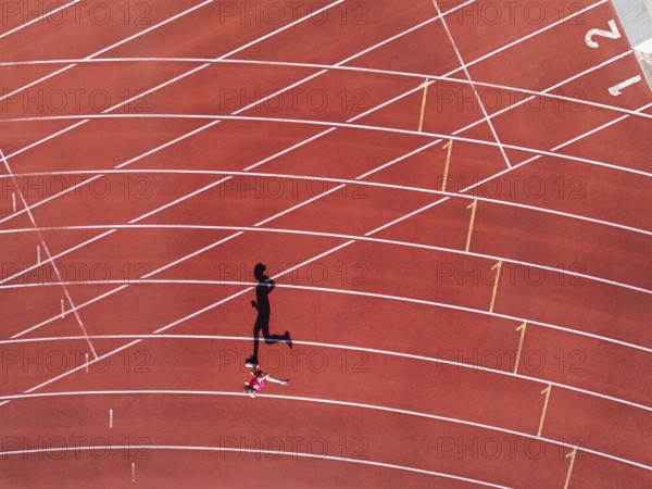 Aerial view of a female individual (runner) on a red athletics track (tartan track) with shadow as a silhouette