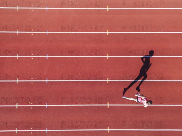 Aerial view of a female individual (runner) on a red athletics track (tartan track) with shadow as a silhouette