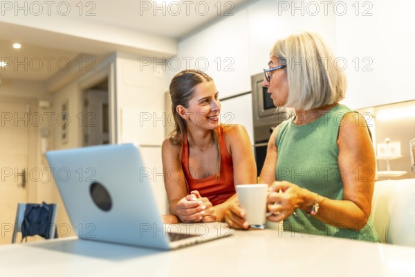 Mother and daughter chatting and smiling while enjoying coffee and browsing a laptop together in a cozy kitchen setting