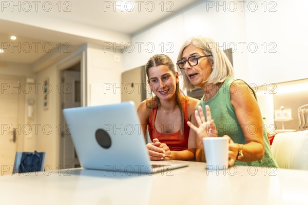 Senior and young women using laptop for a video conference with family, waving and smiling in kitchen