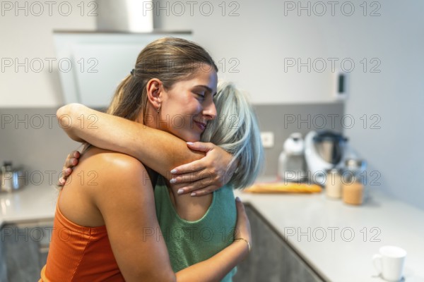 Happy adult daughter is embracing her senior mother in a kitchen, showing love, care, and support