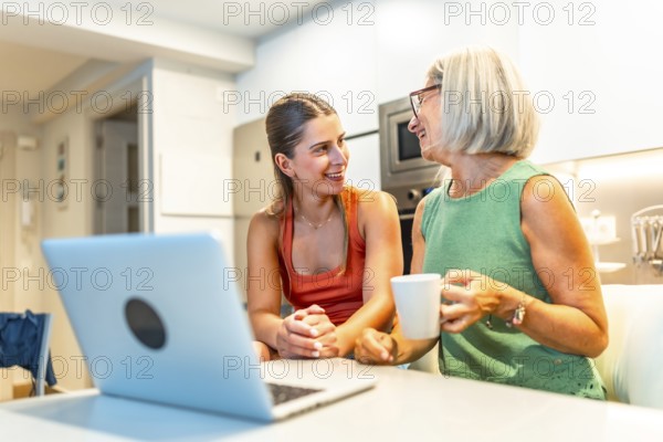 Two women sitting at a kitchen counter, engaging in conversation while using a laptop, one enjoying a warm coffee mug, sharing moments together