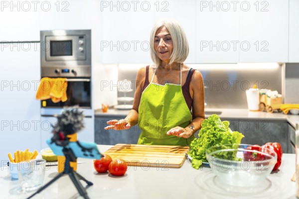 Mature woman in a green apron records a cooking tutorial in a modern kitchen, surrounded by fresh vegetables and kitchen gadgets