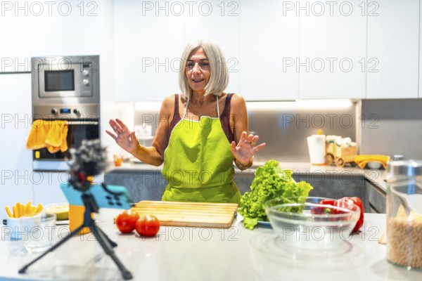 Elderly woman in a green apron recording a cooking tutorial with fresh vegetables, showcasing healthy meal preparation in a contemporary kitchen