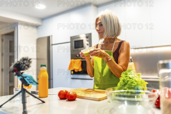 Elderly woman wearing an apron preparing healthy food in her modern kitchen and recording a video recipe with her smartphone