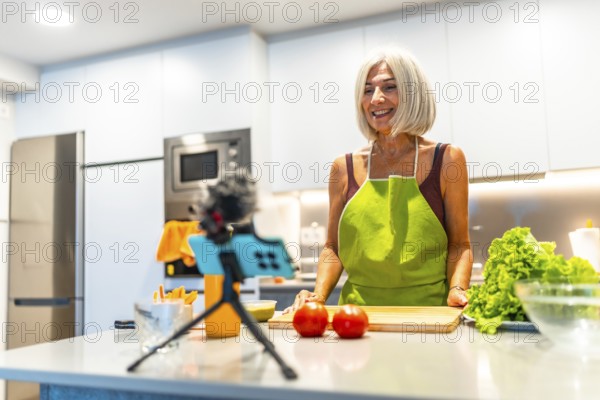 Smiling mature woman wearing apron preparing recipe for her cooking vlog, recording video with smartphone and microphone in kitchen