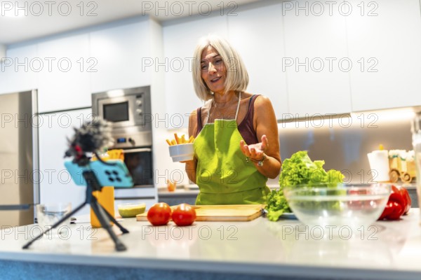 Cheerful mature woman wearing apron holding bowl of carrot sticks while recording video blog about healthy food in kitchen