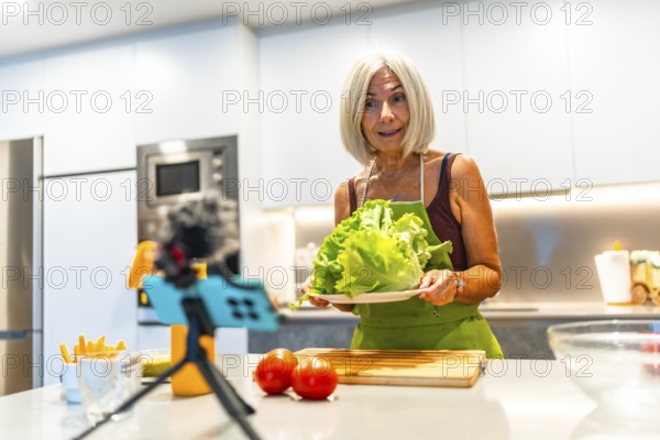 Senior woman creating a cooking video, holding fresh lettuce and surrounded by vegetables in a contemporary kitchen setup