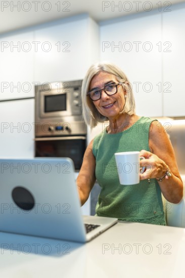 Smiling senior woman enjoying coffee while working on her laptop in a cozy kitchen, embracing a productive and comfortable home office lifestyle