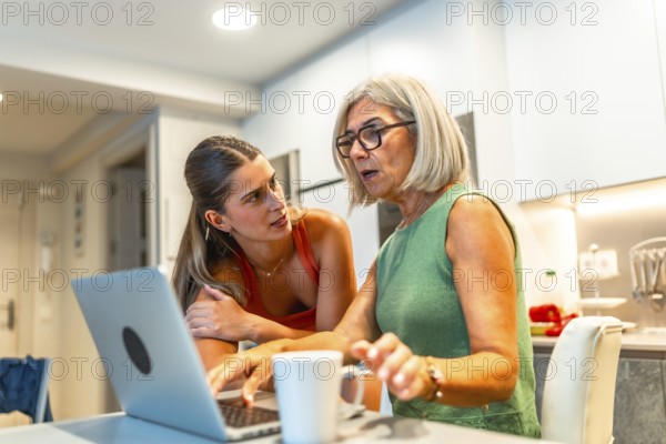 Young woman assisting her senior mother with a laptop in a home kitchen setting, providing technological support and guidance