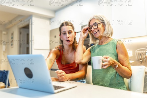 Happy mother and daughter enjoying online streaming content on a laptop while having a coffee in a modern kitchen