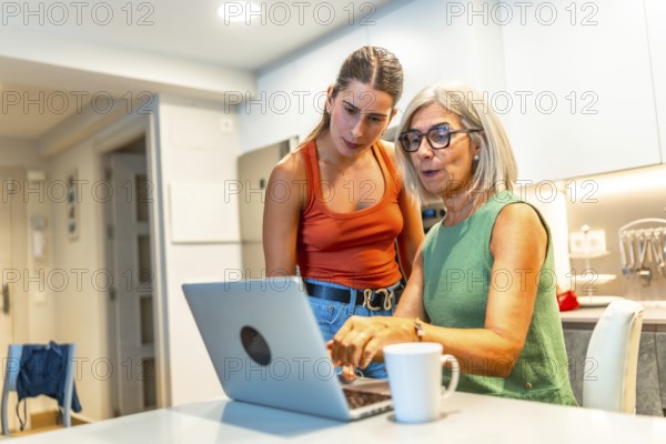Senior woman navigating a laptop with help from a younger woman in a cozy kitchen, fostering family connection and digital skills