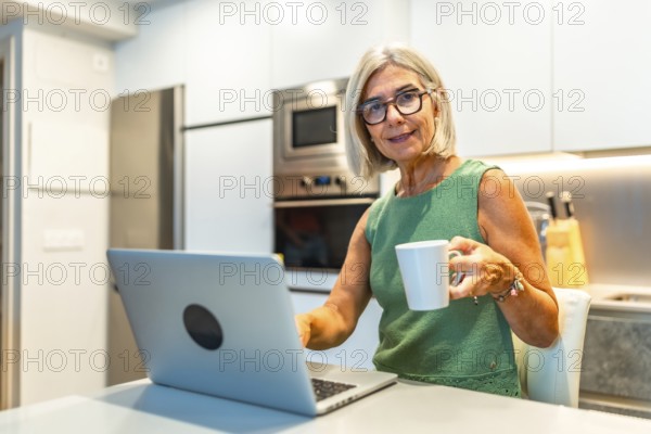 Smiling senior freelancer woman enjoying a cup of coffee while working on her laptop in her modern kitchen