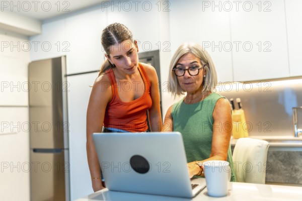 Senior woman and young woman using laptop in kitchen are looking at screen and working on a project