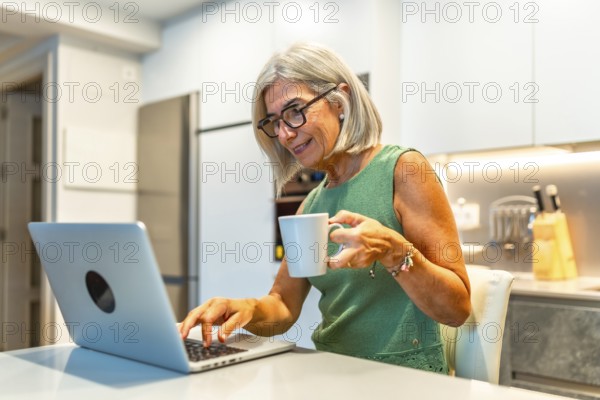 Smiling senior woman enjoying coffee while working on a laptop, embracing the comfort of her home kitchen during remote work hours