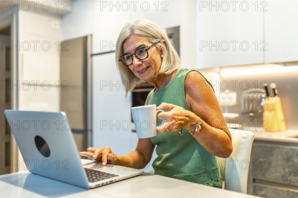 Smiling senior woman using laptop and drinking coffee in modern kitchen, enjoying work from home or retirement