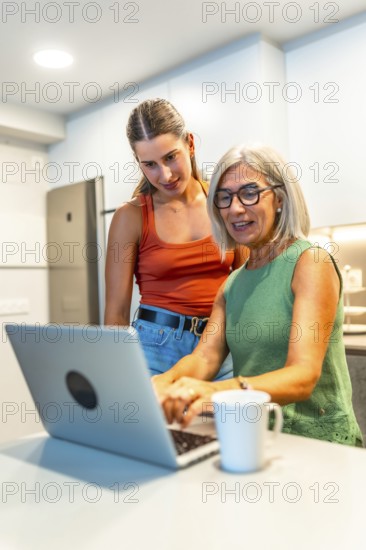 Senior woman using a laptop with guidance from a young woman in a modern kitchen, sharing knowledge and building skills together