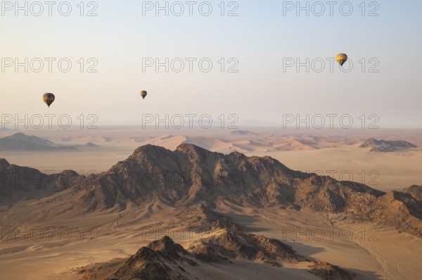 Three hot-air balloons above an arid plain and isolated mountain ridges at the edge of the Namib Desert. Aerial view from another balloon. Namibia