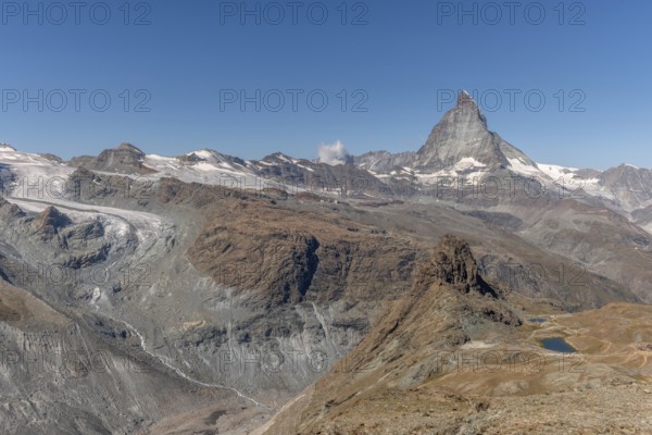 The towering peak of the Matterhorn dominates the clear sky, surrounded by rocky terrain and patches of snow. The barren landscape reveals the natural beauty of the Swiss Alps. Zermatt, Valais